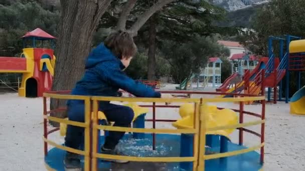 Um menino está montando um carrossel no playground . — Vídeo de Stock