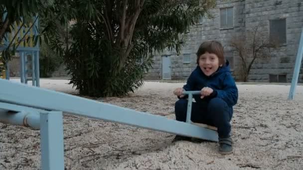Маленький мальчик катается на качелях по детской площадке зимой. Больше никого. . — стоковое видео