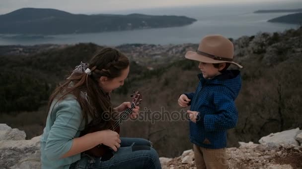 Frau spielt Gitarre und kleiner Junge an bedecktem Tag auf Klippe. — Stockvideo