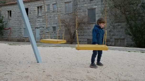Little boy standing near swing in playground outdoors. — Stock Video
