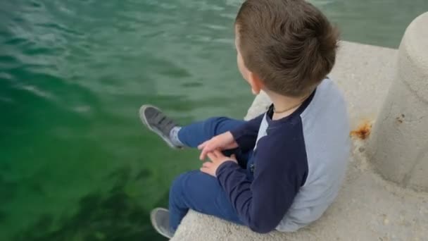 Jongetje bungelt zijn benen vergadering van de kant een pier. — Stockvideo