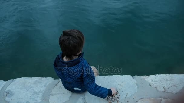 Petit garçon assis sur la jetée jette des pierres dans l'eau à l'extérieur . — Video