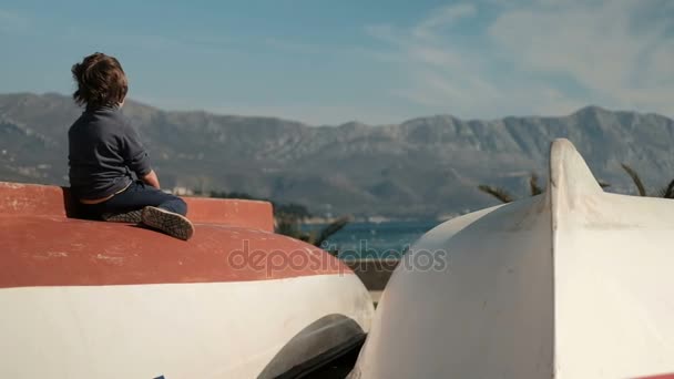 Little boy sitting on boat, looking far near sea outdoors. — Stock Video