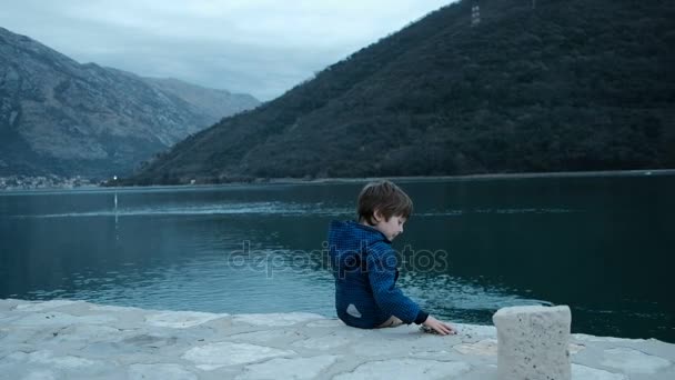 Kleiner Junge sitzt am Ufer und wirft Steine ins Wasser. — Stockvideo
