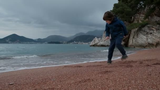 Pequeño niño juega con olas en la orilla del mar al aire libre . — Vídeos de Stock