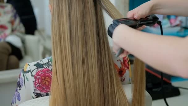 Cabeleireiro endireita o cabelo da mulher sentada com alisador dentro de casa . — Vídeo de Stock