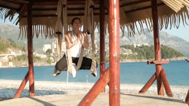 Een vrouw zit in een hangmat in een lotus houding op een zonovergoten strand aan zee. — Stockvideo