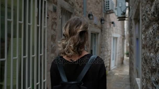 Kvinna med lockigt hår och en ryggsäck som går längs den smala gatan. — Stockvideo