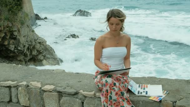 Ung kvinna sitter på havsstranden och målar med akvarell utanför. — Stockvideo
