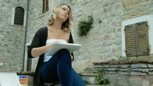 Young woman sits on stones of old city and paints with watercolors. Attractive blond female. — Stock Video