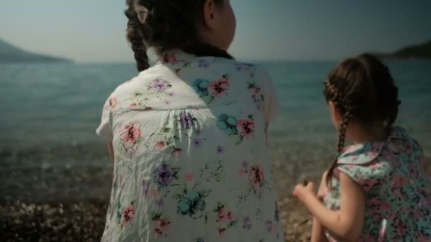 Duas garotas se sentam na costa e jogam pedras na água . — Vídeo de Stock