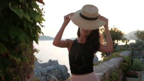 魅力的な女性は岩の多い海岸につば広の帽子を着ています。. — ストック動画