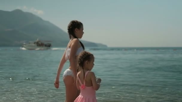 Μικρά κορίτσια στέκονται στην παραλία και να ρίξει πέτρες στο νερό στο ηλιόλουστο καιρό. — Αρχείο Βίντεο