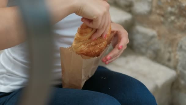 Ung kvinna håller färska bulle i händer sitter på bänken utanför. — Stockvideo