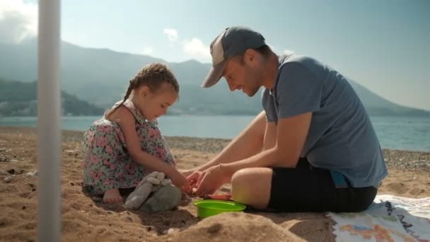 Young man with daughter sits on beach in summer day. — Stock Video