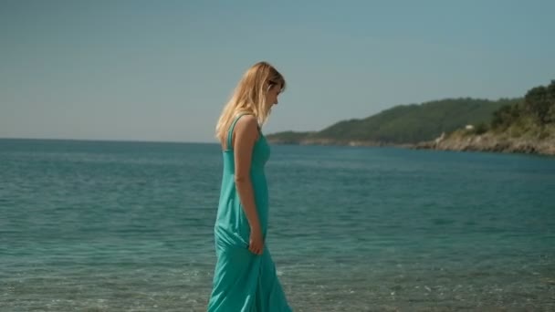 Una mujer en un largo vestido azul pasea sola por la playa . — Vídeos de Stock