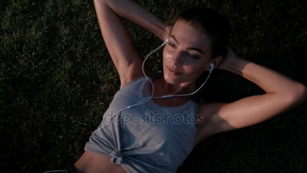 Woman lying on the lawn swinging the press in the headphones. — Stock Video
