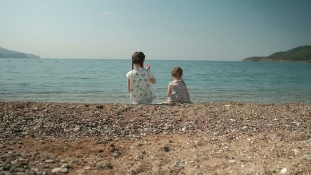Deux filles s'assoient sur le rivage et jettent des pierres dans l'eau en été en plein air . — Video