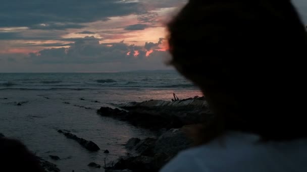 Pareja joven caminando por la noche sobre rocas cerca de la orilla contra un atardecer rojo . — Vídeos de Stock