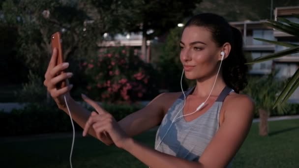 Hermosa mujer con auriculares hace clic en el teléfono en frente de un parque . — Vídeos de Stock