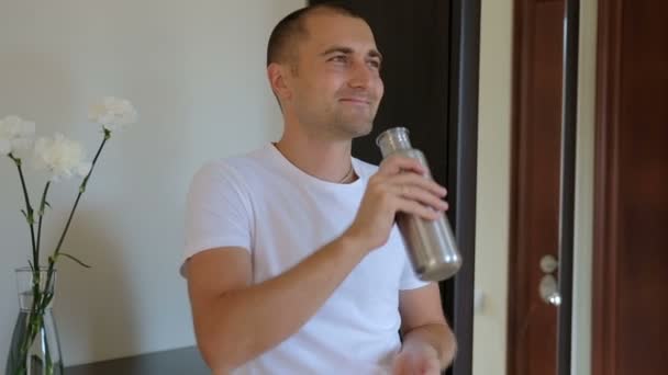 Homem sem barba bebe de uma garrafa térmica de metal e sorri no quarto . — Vídeo de Stock