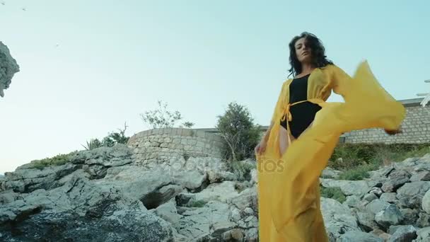 Femme debout sur des rochers agitant un ourlet d'une longue tenue jaune . — Video