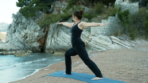 Frau posiert als Kriegerin im Yoga vor dem Hintergrund von Meeresfelsen — Stockvideo