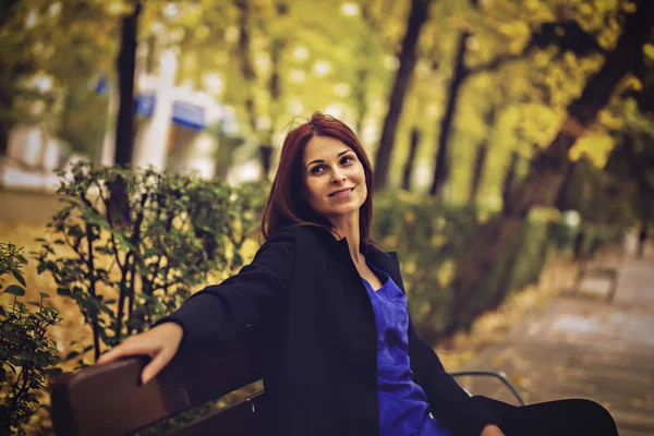 Una mujer hermosa y elegante en el banco en el parque de otoño — Foto de Stock