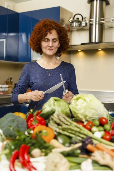 Žena zostření nůž, s hromadou zeleniny před on — Stock fotografie