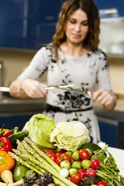 Žena zostření nůž, s hromadou zeleniny před on — Stock fotografie