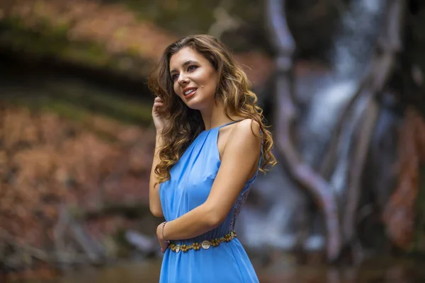 En vacker kvinna i blå klänning nära ett vattenfall på höstdagen — Stockfoto