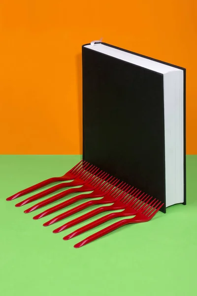 Stillleben mit schwarzem Buch und roten Gabeln auf farbigem Hintergrund — Stockfoto