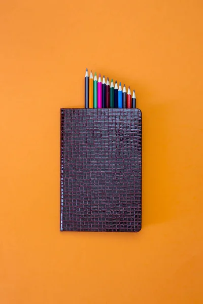 Closed notebook with a set of colored pencils on an orange background, top view