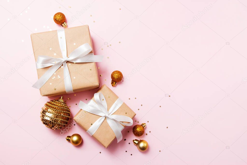 Christmas greeting card composition. Craft paper gift with white ribbon on pink background with christmas golden balls and confetti star. Flat lay, top view, trendy style.