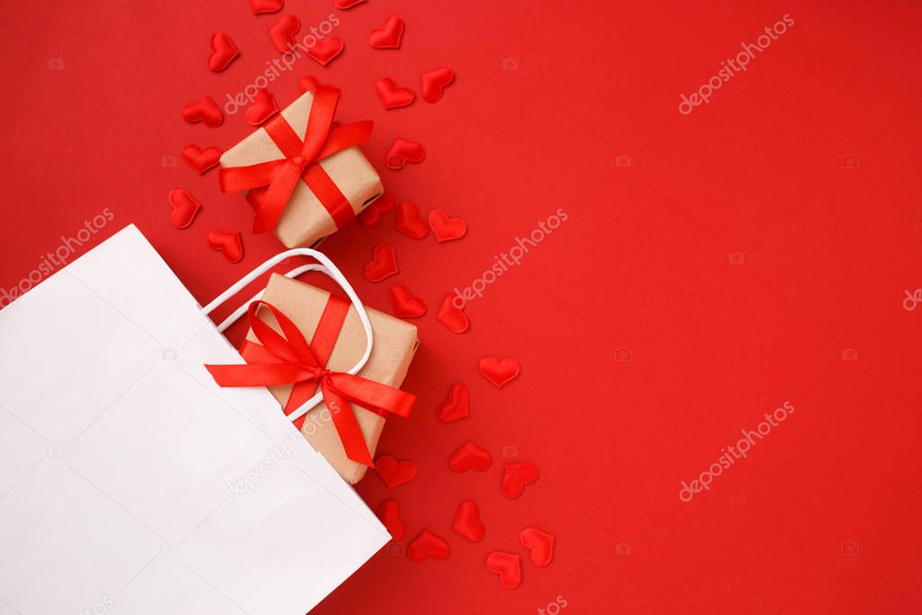 White shopping paper bag with red hearts confetti and gifts on red background. Valentine day concept. Top view, flat lay