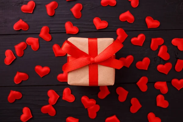 Presente con fiocco rosso su fondo nero in legno con coriandoli a cuore. Stile flat lay. Concetto di San Valentino. Vista dall'alto, piatta — Foto Stock