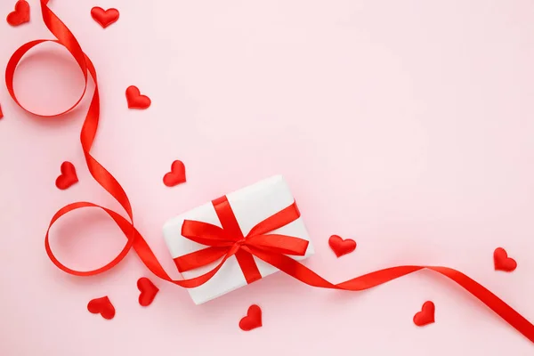 Kırmızı kurdeleli hediye kutusu ve pembe pastel arka planda konfeti kalpleri. Sevgililer Günü tebrik kartı konsepti. Üst görünüm, düz görünüm, kopyalama alanı — Stok fotoğraf