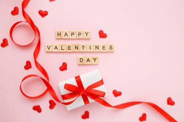 Kırmızı kurdeleli hediye kutusu ve pembe pastel arka planda ahşap harfli konfeti kalpleri. Sevgililer Günü tebrik kartı konsepti. Üst görünüm, düz görünüm, kopyalama alanı