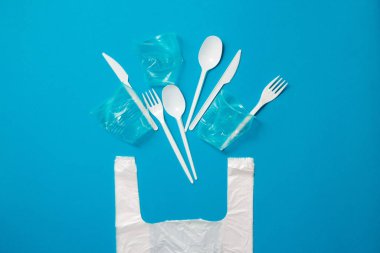 Mavi arka planda beyaz plastik bıçaklar, kaşıklar, çatallar ve torbalar. Tek kullanımlık plastiğe hayır de. Çevresel, kirlilik kavramı.