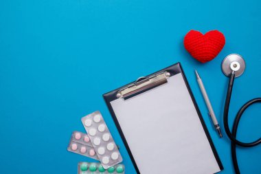 Boş not defterli steteskop, kırmızı kalp ve mavi arka planda haplar. Sağlık hizmetleri konsepti, sağlık çalışanı, kalp sağlık hizmetleri, tıbbi bakım. Düz düzen, üst görünüm