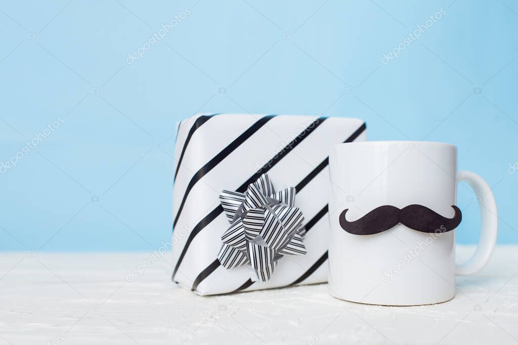 Greeting card fathers day holiday concept. White cup with mustache and gift box on blue pastel background