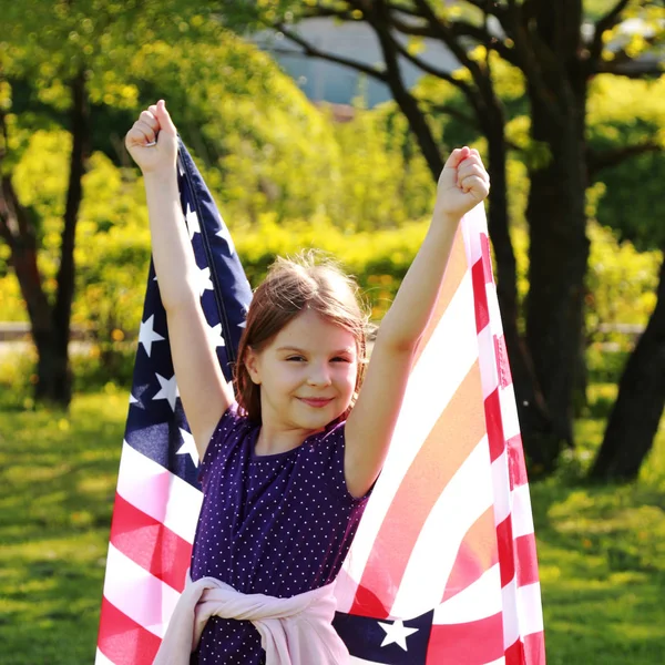 公園で日当たりの良い夏の日の大規模な米国旗を保持している白いドレスを着てうれしそうな美少女 — ストック写真