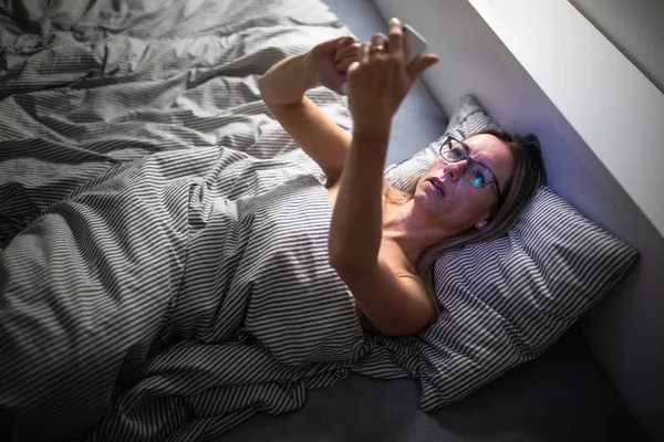 예쁘고 중년의 여성 이 침대에서 핸드폰을 사용하고 있습니다. — 스톡 사진