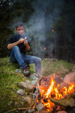 Genç adam kamp ateşinin yanında ağaçtan aletler yapıyor.