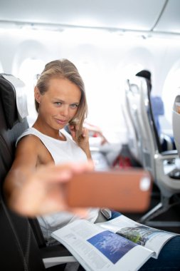 Genç, mutlu bir kadın, pasaportlu bir belgeyle fotoğraf çekiyor. Uçak uçuşu sırasında pencere kenarındaki koltukta oturuyor.