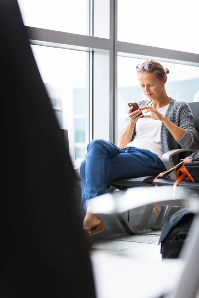 国際空港の出発ゲートで飛行機に搭乗を待っている間彼女の携帯電話を使用してカジュアルな金髪の若い女性 — ストック写真