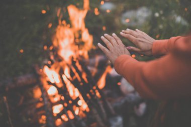 Dışarıda kamp yaparken ateş yakan genç bir kadın, alp yabanda ellerini ateşin yanında ısıtıyor.