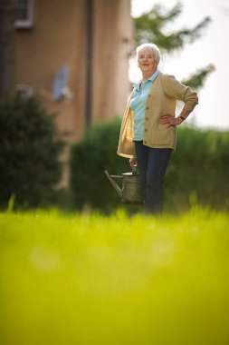 Yaşlı kadın bahçesinde bahçe işleriyle uğraşıyor. Çiçekleri suluyor.