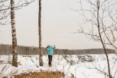 Kışın kış ormanarka üzerinde uçurumun kenarında parlak giysiler içinde kız. Telefonun elinde, telefonda fotoğraf çekmek