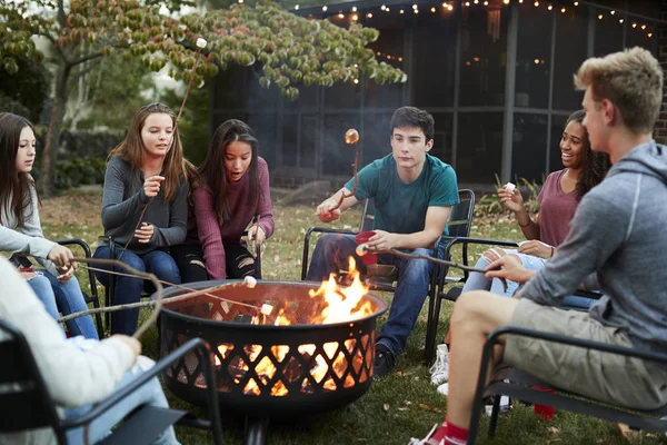 Teenage friends sit round a fire pit toasting marshmallows